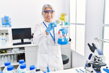 Téléchargez les photos : Femme aux cheveux gris d'âge moyen portant l'uniforme de scientifique versant du liquide sur une éprouvette en laboratoire - en image libre de droit