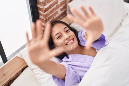 Foto de Joven mujer hispana hermosa acostada en la cama haciendo gesto fotográfico con las manos en el dormitorio - Imagen libre de derechos