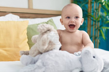 Photo for Adorable caucasian baby sitting on bed with dolls smiling at bedroom - Royalty Free Image