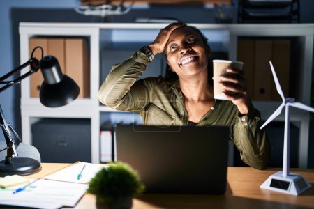 Téléchargez les photos : Femme africaine travaillant à l'aide d'un ordinateur portable la nuit souriant confiant toucher les cheveux avec geste de la main vers le haut, posant attrayant et à la mode - en image libre de droit