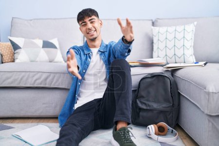 Photo for Young hispanic man sitting on the floor studying for university looking at the camera smiling with open arms for hug. cheerful expression embracing happiness. - Royalty Free Image