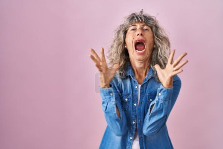 Foto de Mujer de mediana edad de pie sobre fondo rosa loco y loco gritando y gritando con expresión agresiva y los brazos levantados. concepto de frustración. - Imagen libre de derechos