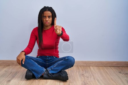 Téléchargez les photos : Young african american with braids sitting on the floor at home pointing with finger up and angry expression, showing no gesture - en image libre de droit