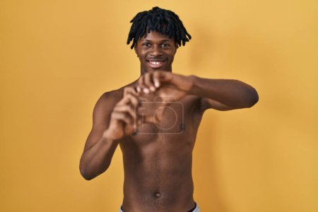Téléchargez les photos : Jeune homme africain avec dreadlocks debout torse nu souriant dans l'amour faisant forme de symbole de coeur avec les mains. concept romantique. - en image libre de droit