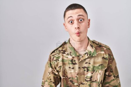 Foto de Joven vestido con camuflaje uniforme del ejército haciendo cara de pez con labios, gesto loco y cómico. expresión divertida. - Imagen libre de derechos