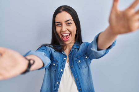Photo for Hispanic woman standing over blue background looking at the camera smiling with open arms for hug. cheerful expression embracing happiness. - Royalty Free Image