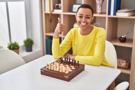 Foto de Mujer afroamericana jugando ajedrez sentado en la mesa sonriendo feliz y positivo, pulgar hacia arriba haciendo excelente y signo de aprobación - Imagen libre de derechos