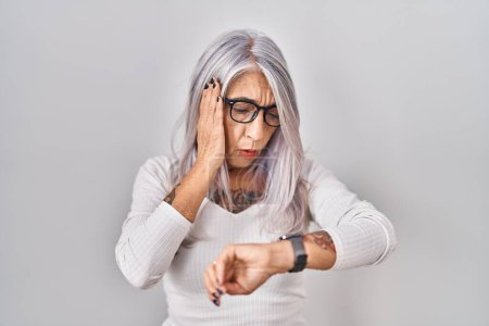 Téléchargez les photos : Femme d'âge moyen avec les cheveux gris debout sur fond blanc regardant le temps de la montre inquiet, peur de se faire tard - en image libre de droit