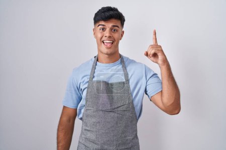 Foto de Hispanic young man wearing apron over white background pointing finger up with successful idea. exited and happy. number one. - Imagen libre de derechos