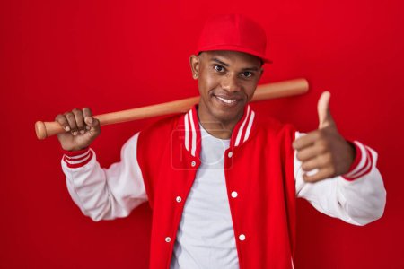 Téléchargez les photos : Young hispanic man playing baseball holding bat approving doing positive gesture with hand, thumbs up smiling and happy for success. winner gesture. - en image libre de droit