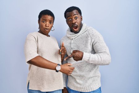 Téléchargez les photos : Jeune couple afro-américain debout sur fond bleu ensemble à la hâte pointant vers regarder le temps, l'impatience, bouleversé et en colère pour retard de délai - en image libre de droit