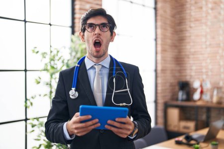 Photo for Young hispanic man wearing doctor stethoscope at the office angry and mad screaming frustrated and furious, shouting with anger looking up. - Royalty Free Image