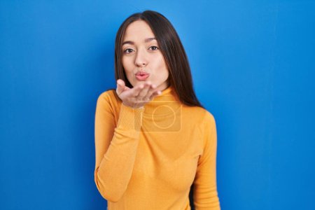 Photo for Young brunette woman standing over blue background looking at the camera blowing a kiss with hand on air being lovely and sexy. love expression. - Royalty Free Image