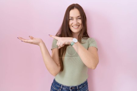 Photo for Beautiful brunette woman standing over pink background amazed and smiling to the camera while presenting with hand and pointing with finger. - Royalty Free Image