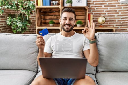 Foto de Hombre hispano guapo haciendo compras en línea sosteniendo tarjeta de crédito haciendo signo bien con los dedos, sonriendo gesto amistoso excelente símbolo - Imagen libre de derechos