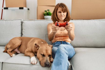Téléchargez les photos : Jeune femme caucasienne jouant jeu vidéo assis sur le canapé avec chien à la maison - en image libre de droit