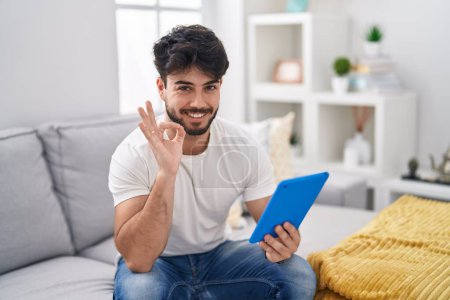 Foto de Hombre hispano con barba usando touchpad sentado en el sofá sonriendo positivo haciendo signo de ok con la mano y los dedos. expresión exitosa. - Imagen libre de derechos