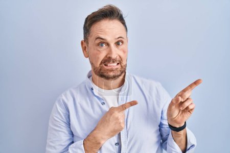 Photo for Middle age caucasian man standing over blue background pointing aside worried and nervous with both hands, concerned and surprised expression - Royalty Free Image