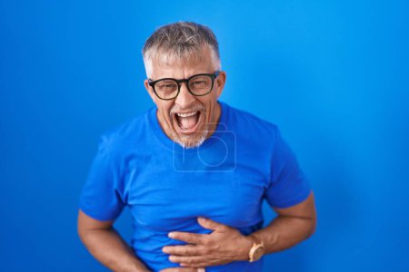 Téléchargez les photos : Hispanic man with grey hair standing over blue background smiling and laughing hard out loud because funny crazy joke with hands on body. - en image libre de droit