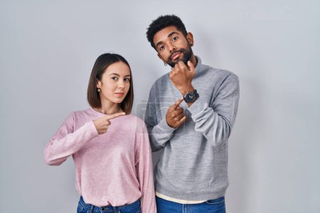 Téléchargez les photos : Young hispanic couple standing together in hurry pointing to watch time, impatience, looking at the camera with relaxed expression - en image libre de droit