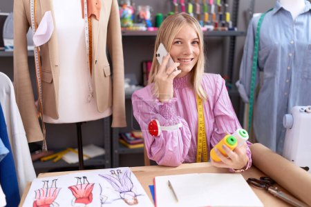 Téléchargez les photos : Young blonde woman tailor talking on smartphone holding thread at tailor shop - en image libre de droit