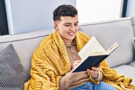 Téléchargez les photos : Jeune homme non binaire livre de lecture assis sur le canapé à la maison - en image libre de droit