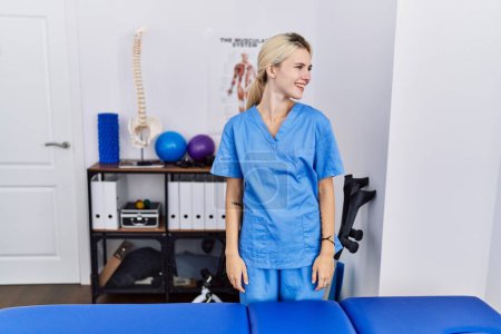Photo for Young physiotherapist woman working at pain recovery clinic looking away to side with smile on face, natural expression. laughing confident. - Royalty Free Image