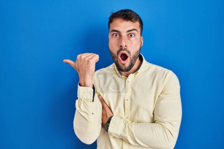 Photo for Handsome hispanic man standing over blue background surprised pointing with hand finger to the side, open mouth amazed expression. - Royalty Free Image