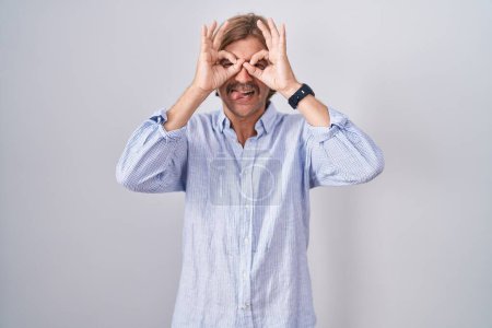 Foto de Caucasian man with mustache standing over white background doing ok gesture like binoculars sticking tongue out, eyes looking through fingers. crazy expression. - Imagen libre de derechos