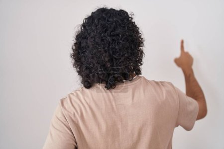 Foto de Hombre hispano con el pelo rizado de pie sobre fondo blanco posando hacia atrás apuntando hacia adelante con la mano del dedo - Imagen libre de derechos