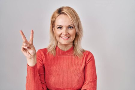 Photo for Blonde woman standing over isolated background smiling with happy face winking at the camera doing victory sign with fingers. number two. - Royalty Free Image