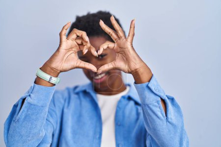 Photo for African american woman standing over blue background doing heart shape with hand and fingers smiling looking through sign - Royalty Free Image
