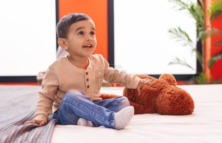Téléchargez les photos : Adorable bambin hispanique souriant confiant assis sur le lit dans la chambre - en image libre de droit