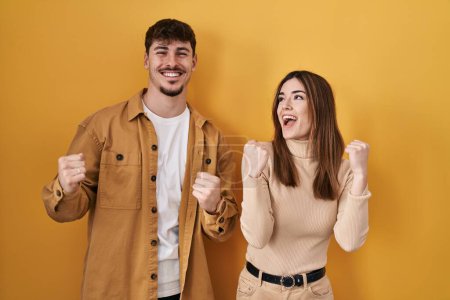 Photo for Young hispanic couple standing over yellow background celebrating surprised and amazed for success with arms raised and eyes closed. winner concept. - Royalty Free Image