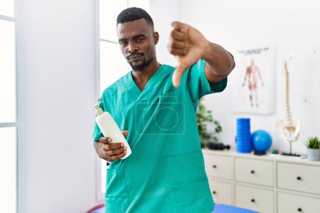 Foto de Young african physiotherapist man holding massage body lotion with angry face, negative sign showing dislike with thumbs down, rejection concept - Imagen libre de derechos