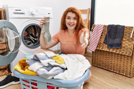 Foto de Young redhead woman putting dirty laundry into washing machine smiling friendly offering handshake as greeting and welcoming. successful business. - Imagen libre de derechos