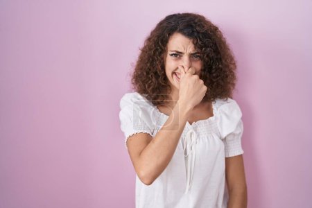Foto de Hispanic woman with curly hair standing over pink background smelling something stinky and disgusting, intolerable smell, holding breath with fingers on nose. bad smell - Imagen libre de derechos