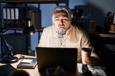 Téléchargez les photos : Homme d'âge moyen avec les cheveux gris travaillant au bureau la nuit en regardant la caméra souffler un baiser sur l'air étant belle et sexy. expression d'amour. - en image libre de droit