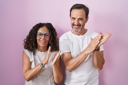 Foto de Mediana edad pareja hispana juntos sobre rosa fondo aplaudiendo y aplaudiendo feliz y alegre, sonriendo orgullosas manos juntos - Imagen libre de derechos