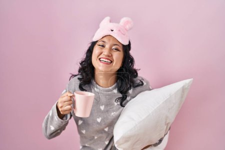 Foto de Young asian woman wearing sleep mask and pajama hugging pillow drinking coffee smiling and laughing hard out loud because funny crazy joke. - Imagen libre de derechos