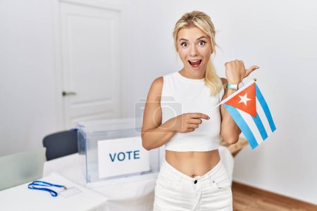 Foto de Mujer caucásica joven en las elecciones de campaña política sosteniendo la bandera de Cuba señalando el pulgar hacia el lado sonriendo feliz con la boca abierta - Imagen libre de derechos