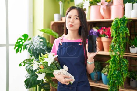 Foto de Young chinese woman working at florist shop showing smartphone screen looking at the camera blowing a kiss being lovely and sexy. love expression. - Imagen libre de derechos