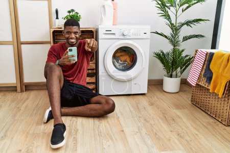 Foto de Joven hombre afroamericano usando smartphone esperando a que la lavadora apunte a usted y a la cámara con los dedos, sonriendo positiva y alegre - Imagen libre de derechos
