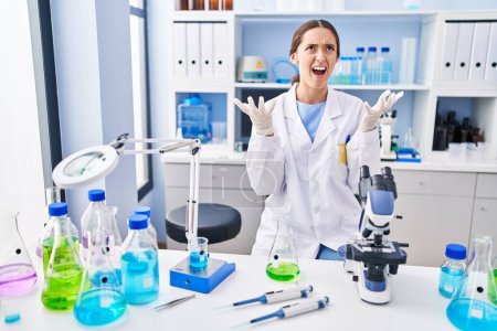 Foto de Young brunette woman working at scientist laboratory crazy and mad shouting and yelling with aggressive expression and arms raised. frustration concept. - Imagen libre de derechos