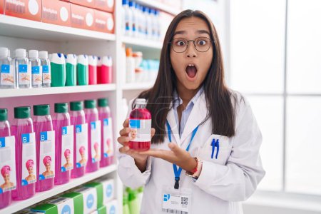 Photo for Young hispanic woman working at pharmacy drugstore holding syrup afraid and shocked with surprise and amazed expression, fear and excited face. - Royalty Free Image