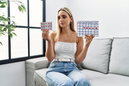 Photo for Young blonde woman holding period calendar and pills at home - Royalty Free Image