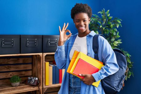 Photo for African american woman wearing student backpack and holding books doing ok sign with fingers, smiling friendly gesturing excellent symbol - Royalty Free Image