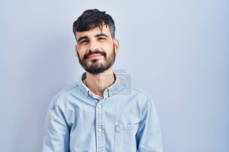 Photo for Young hispanic man with beard standing over blue background with hands together and crossed fingers smiling relaxed and cheerful. success and optimistic - Royalty Free Image