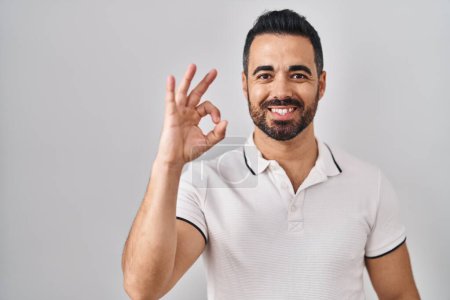 Foto de Joven hombre hispano con barba vistiendo ropa casual sobre fondo blanco sonriendo positivo haciendo signo de ok con la mano y los dedos. expresión exitosa. - Imagen libre de derechos