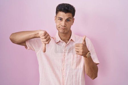 Foto de Young hispanic man standing over pink background doing thumbs up and down, disagreement and agreement expression. crazy conflict - Imagen libre de derechos